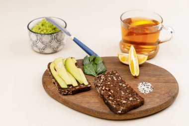 Avokado tostu için malzeme içeren ahşap tahta. Avokado dilimli koyu çavdar ekmeği, taze fesleğen yaprağı, limon dilimleri ve susam tohumları. Kase ve çayın içinde guacamole 'nin yanında. Sağlıklı kahvaltı kavramı