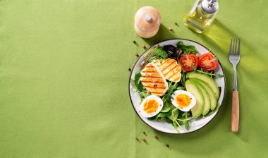 Sağlıklı keto diyeti kahvaltısı: haşlanmış yumurta, avokado dilimleri, ızgara halloumi peyniri, salata yaprakları. Yeşil arka plan. en üstteki görünüm