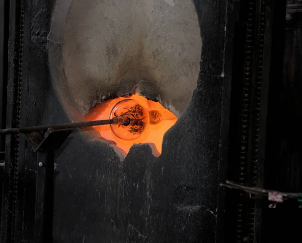 Long metal rod holds unit of glass in a specialized fiery oven in a glass blowers workshop and creates an eerie image when seen close up..