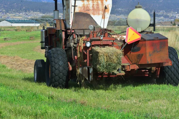 Agriculture: hay baler moves along a row of raked hay, scoops it up, compresses it, ties it tight into a bale. It will then drop it and take up more loose hay from the field.