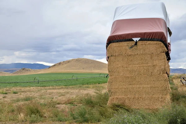 Hay storage: Large stack of square hay bales in a field with protective tarp on top, distant hills and cloudy sky