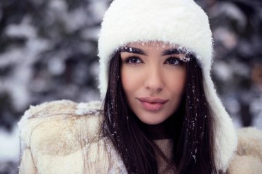 Beyaz kürklü ve beyaz şapkalı uzun saçlı güzel esmer kız otelde eğleniyor. Dış mekan fotoğrafçılığını kapat.