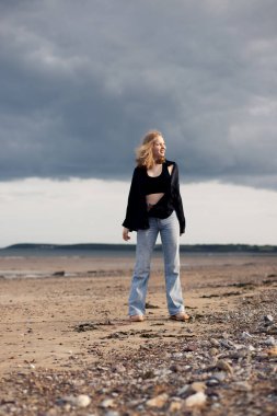 Siyah ipek gömlek ve kot pantolon giymiş genç sarışın bir kız kumsalda yürüyor ve bulutlu havanın tadını çıkarıyor.