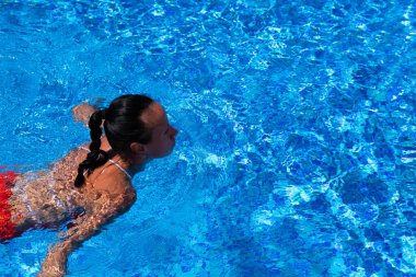 Bronzlaşmış bir kızın üst görüntüsü, kırmızı mayo giymiş bir model, havuzun mavi sularında yüzüyor..