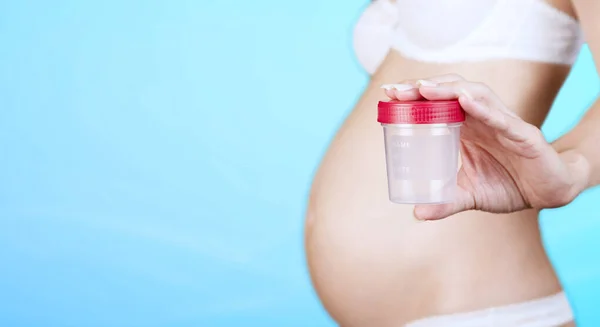 İç çamaşırlı hamile bir beyaz kadın elinde idrar toplamak için plastik bir kap, mavi arka planda kırmızı başlıklı bir kavanoz tutuyor. Yakın plan. Uzayı kopyala Yüksek kalite fotoğraf