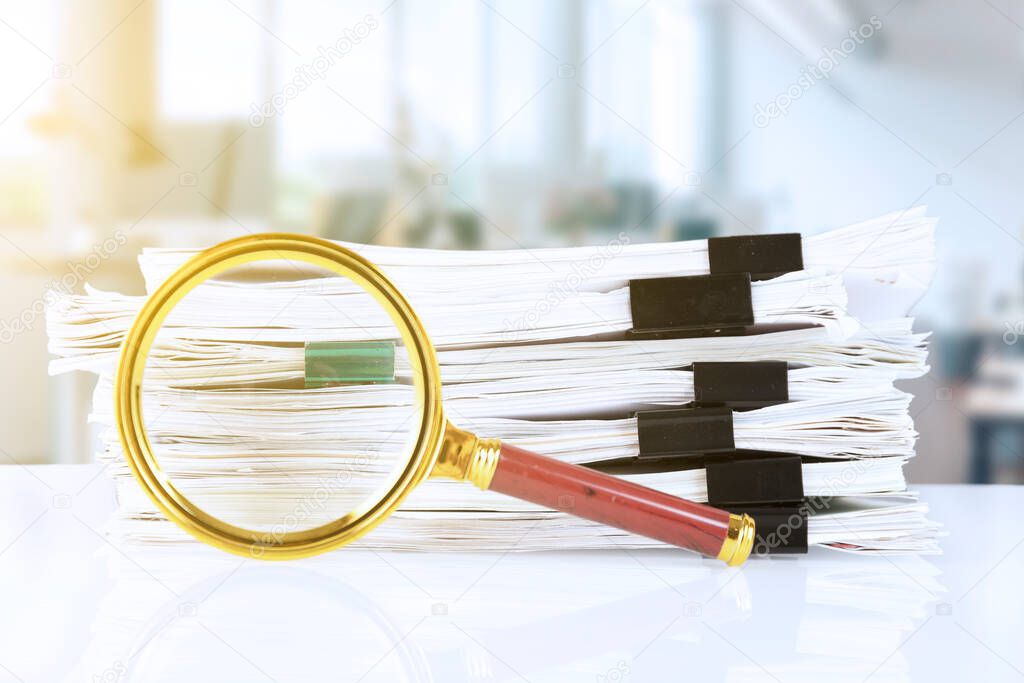 stack of paper documents with a magnifying glass on an office desk, against a blurred office background. Business and search concept. High quality photo