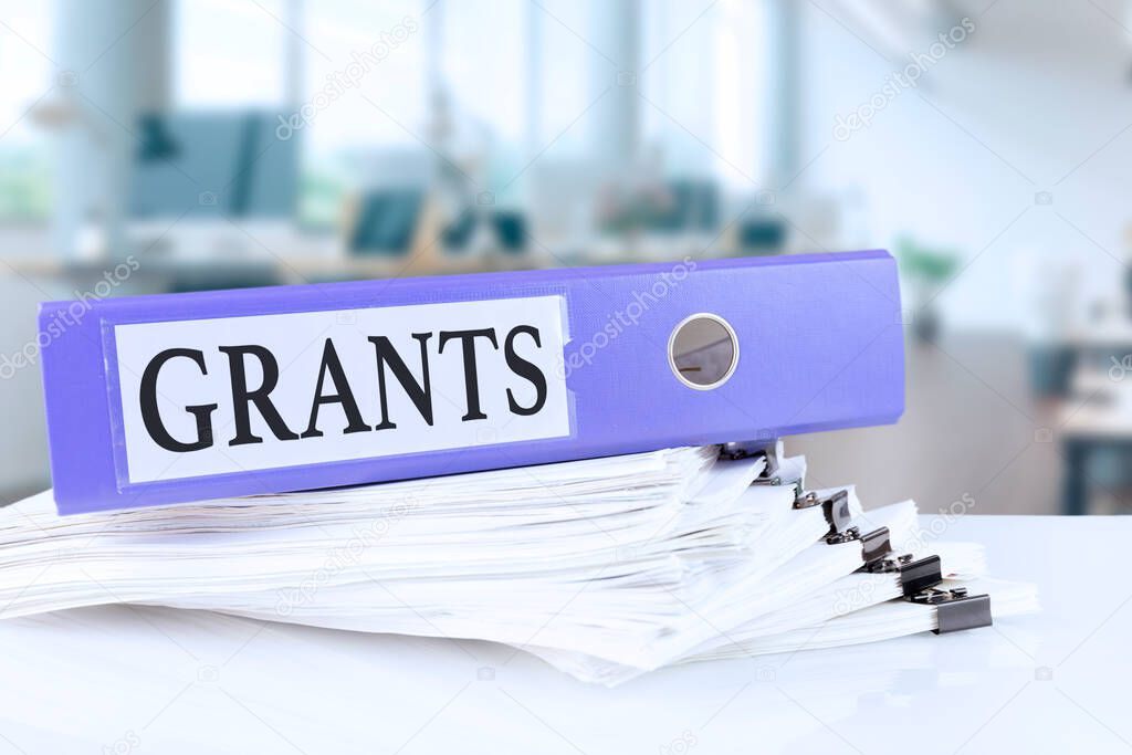 Grants, the term is written on a folder of documents in trendy purple, lying on a stack of documents on an office table against the backdrop of an office with a soft blurred background. High quality photo