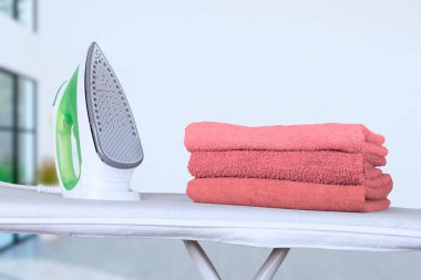 Elektrikli ütü bir ütü masasının üzerinde duruyor ve pembe banyo havlularını evin arka planına doğru katlıyor. Bulanık arka plan, yumuşak odaklanma. Yüksek kalite fotoğraf