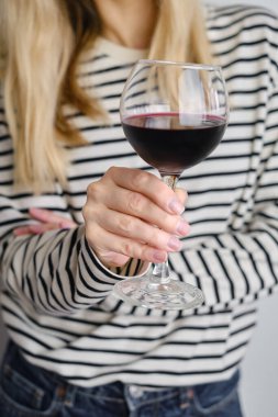 A glass of red wine in the hands of a girl in a striped jacket against the background of a white wall. Wine tasting at the bar. Party, drinks, holidays and celebration concept. Glass close-up