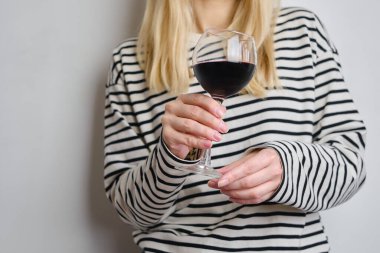 A glass of red wine in the hands of a girl in a striped jacket against the background of a white wall. Wine tasting at the bar. Party, drinks, holidays and celebration concept. Glass close-up