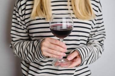 A glass of red wine in the hands of a girl in a striped jacket against the background of a white wall. Wine tasting at the bar. Party, drinks, holidays and celebration concept. Glass close-up