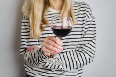A glass of red wine in the hands of a girl in a striped jacket against the background of a white wall. Wine tasting at the bar. Party, drinks, holidays and celebration concept. Glass close-up