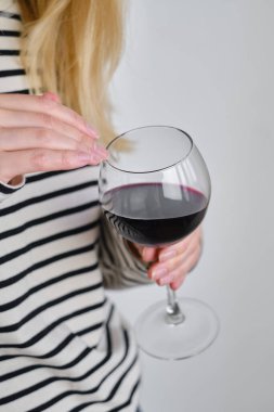 A glass of red wine in the hands of a girl in a striped jacket against the background of a white wall. Wine tasting at the bar. Party, drinks, holidays and celebration concept. Glass close-up