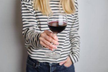 A glass of red wine in the hands of a girl in a striped jacket against the background of a white wall. Wine tasting at the bar. Party, drinks, holidays and celebration concept. Glass close-up