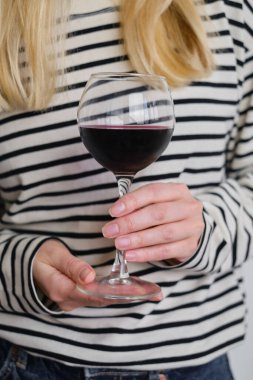 A glass of red wine in the hands of a girl in a striped jacket against the background of a white wall. Wine tasting at the bar. Party, drinks, holidays and celebration concept. Glass close-up