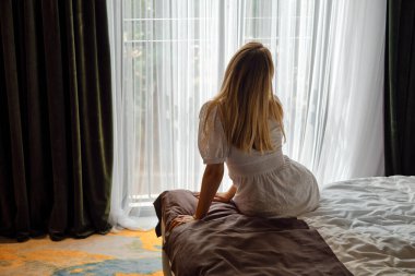 A girl in a white dress stretches after getting out of bed and looking out the window. Morning rituals for a vigorous workday. Health and relaxation concept