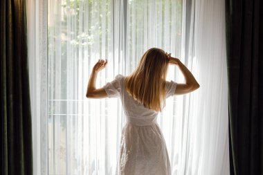 A girl in a white dress stretches after getting out of bed and looking out the window. Morning rituals for a vigorous workday. Health and relaxation concept