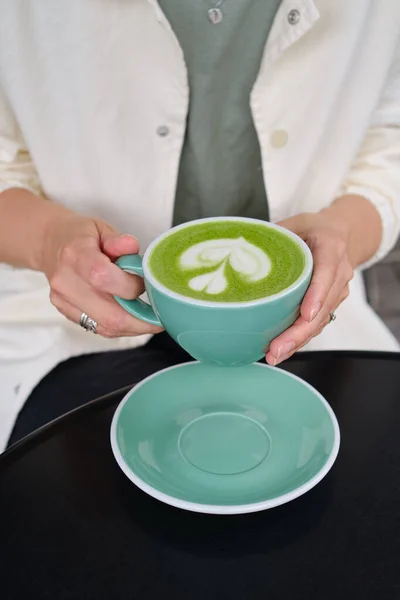 Woman with a cup of matcha latte sits on the summer terrace of a cafe. Delicious green matcha tea in a green cup close-up. Japanese hot milky beverage. Healthy Matcha Tea Made with Organic Ingredients