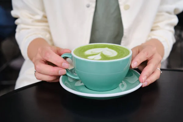 Woman with a cup of matcha latte sits on the summer terrace of a cafe. Delicious green matcha tea in a green cup close-up. Japanese hot milky beverage. Healthy Matcha Tea Made with Organic Ingredients