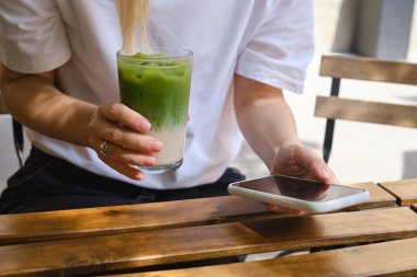 Girl drinks an ice matcha latte and uses a smartphone. A woman is holding a mobile phone, sending a text message or using an application on her mobile phone. Delicious green organic matcha tea