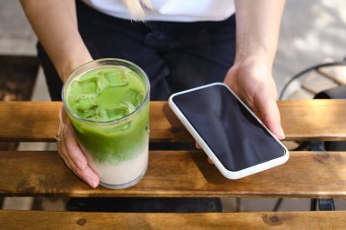 Girl drinks an ice matcha latte and uses a smartphone. A woman is holding a mobile phone, sending a text message or using an application on her mobile phone. Delicious green organic matcha tea