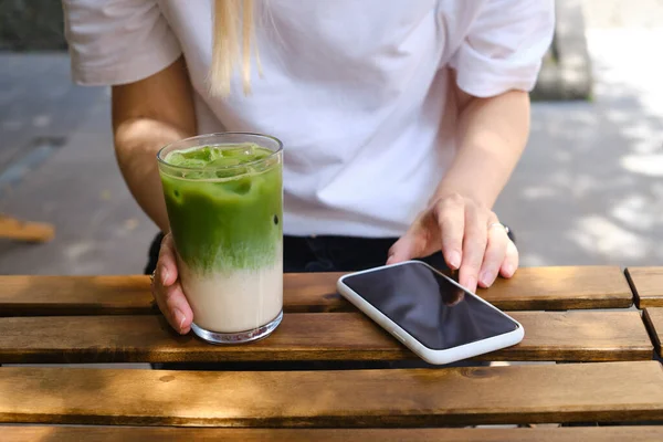 Girl drinks an ice matcha latte and uses a smartphone. A woman is holding a mobile phone, sending a text message or using an application on her mobile phone. Delicious green organic matcha tea
