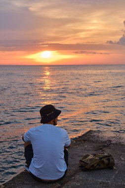 A young man sits by the sea and looks at the dawn. Male on travels. Amazing picturesque outdoor view. Beautiful dawn on the seashore. Healthy Lifestyle, Leisure Activities. Travel and Exploration