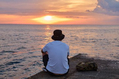 A young man sits by the sea and looks at the dawn. Male on travels. Amazing picturesque outdoor view. Beautiful dawn on the seashore. Healthy Lifestyle, Leisure Activities. Travel and Exploration
