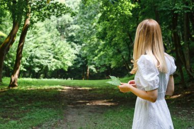 Beautiful woman in a white dress walks along the green path in a picturesque park. Outdoor activities. Happy holidays outside the city. Summer walk in nature. Enjoy the weather and fresh air