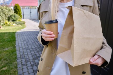 A paper glass and a craft bag in the hands of a girl. A glass for coffee and hot drinks to go. Food delivery service. Paper bag close up. Blank space mockup for design, lettering or advertising