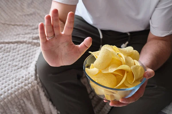 A man eats crispy potato chips from a transparent bowl on the couch. Quick snack. Calories and diet