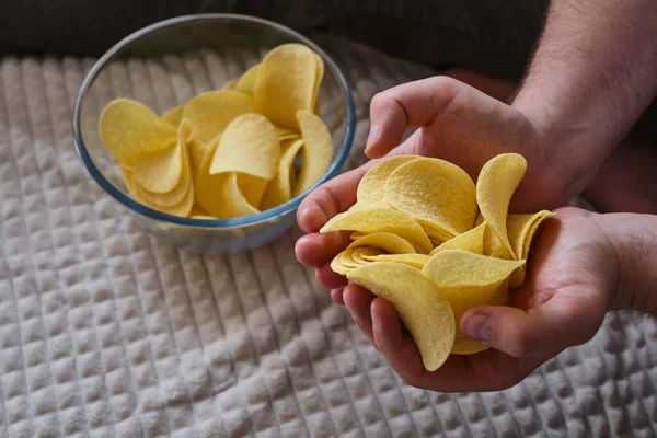 A man eats crispy potato chips from a transparent bowl on the couch. Quick snack. Calories and diet