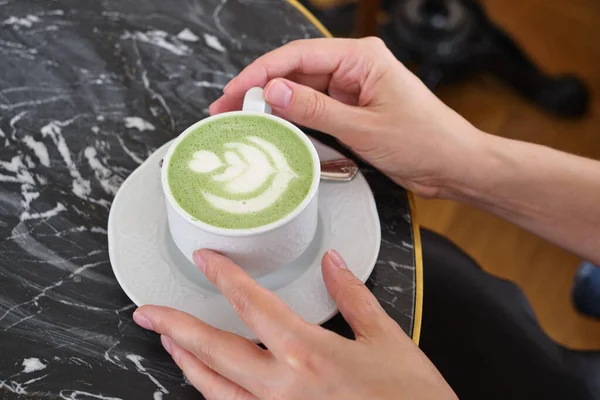Woman with a cup of matcha latte sits in the cafe. Delicious green matcha tea in a green cup close-up. Japanese hot milky beverage. Healthy Matcha Tea Made with Organic Ingredients