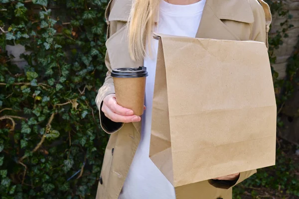 A paper glass and a craft bag in the hands of a girl. A glass for coffee and hot drinks to go. Food delivery service. Paper bag close up. Blank space mockup for design, lettering or advertising