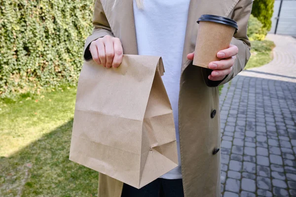 A paper glass and a craft bag in the hands of a girl. A glass for coffee and hot drinks to go. Food delivery service. Paper bag close up. Blank space mockup for design, lettering or advertising