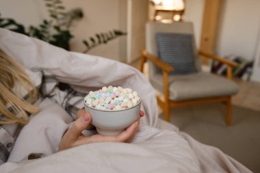 A girl in pajamas sits on the windowsill and holds a cup of cocoa with marshmallows. Cozy winter days at home. Quarantine time
