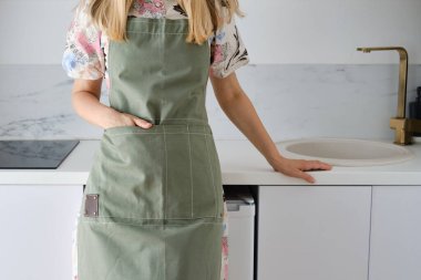 Mutfak önlüklü bir kadın modern bir mutfakta duruyor. Üniformayla evde yemek pişirmek, koruyucu giysi. Yeşil kumaş önlük, günlük kıyafetler. Önlüklü kız yemek yapmaya hazırlanıyordu.