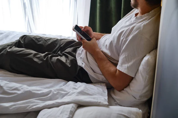 A man holds a smartphone in his hands in bed and checks a social network account before going to bed or after waking up. Gadget addiction. Man sending a text message on a mobile phone