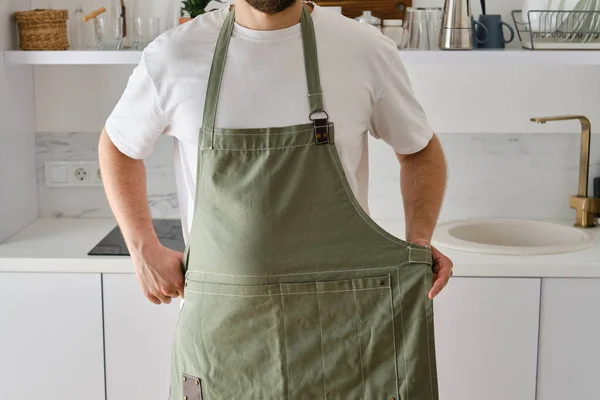 A man in a kitchen apron stands in a modern kitchen. Cooking at home in uniform, protection apparel. Green fabric apron, casual clothing. A man in an apron prepares to prepare a meal
