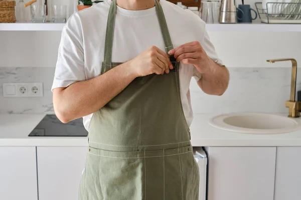 A man in a kitchen apron stands in a modern kitchen. Cooking at home in uniform, protection apparel. Green fabric apron, casual clothing. A man in an apron prepares to prepare a meal