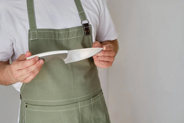 A man in a kitchen apron and a knife stands in a modern kitchen. Cooking at home in uniform, protection apparel. Green fabric apron, casual wear. A man in an apron prepares to prepare a meal