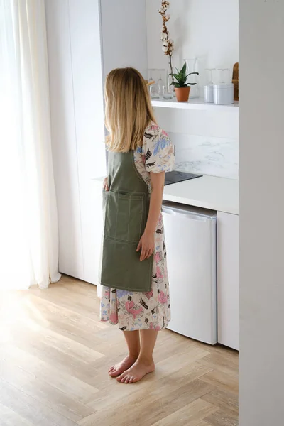 A woman in a kitchen apron stands in a modern kitchen. Cooking at home in uniform, protection apparel. Green fabric apron, casual clothing. The girl in the apron got ready to cook