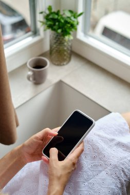 Pencerenin yanında elinde bir fincan sabah kahvesi olan bir kız modern bir akıllı telefon tutuyor. Bir kadın cep telefonu tutuyor, mesaj gönderiyor ya da cep telefonunda bir uygulama kullanıyor