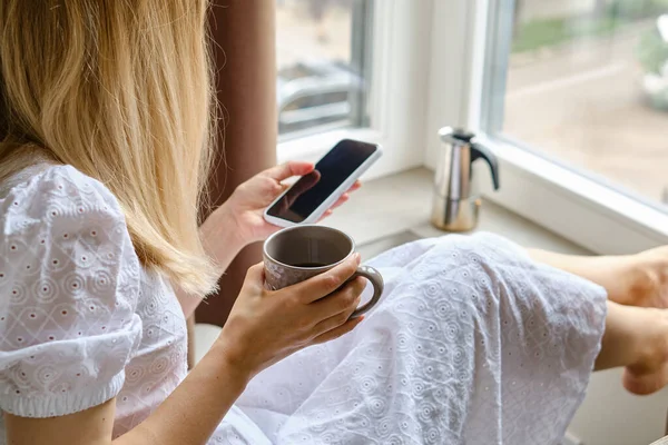 A girl near the window with a cup of morning coffee is holding a modern smartphone. A woman is holding a mobile phone, sending a text message or using an application on her mobile phone