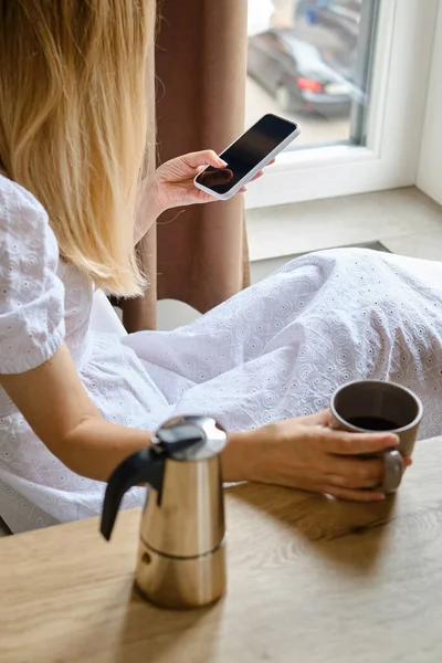 A girl near the window with a cup of morning coffee is holding a modern smartphone. A woman is holding a mobile phone, sending a text message or using an application on her mobile phone