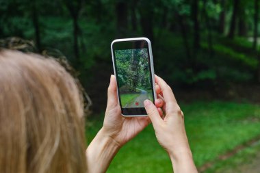 Kız elinde bir cep telefonu tutuyor ve sosyal ağlarda fotoğrafları paylaşmak için parkta doğanın fotoğraflarını çekiyor. Telefon ekranına odaklan. Akıllı telefonunla anılar yarat. Cep telefonunu kapat.