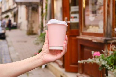 Bir kadının elinde kahve olan pembe kağıt bardak. Şehirde kahve içmenin zamanı geldi. Şehrin arka planında tek kullanımlık kağıt bardak var. Metin ve düzen için boş alan. Kahve konsepti.