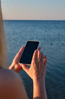 Kız elinde bir cep telefonu tutuyor ve şafakta denizin fotoğraflarını çekiyor. Telefon ekranına odaklan. Akıllı telefonunla anılar yarat. Cep telefonunu kapat.