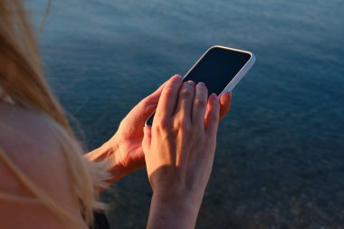 Kız elinde bir cep telefonu tutuyor ve şafakta denizin fotoğraflarını çekiyor. Telefon ekranına odaklan. Akıllı telefonunla anılar yarat. Cep telefonunu kapat.