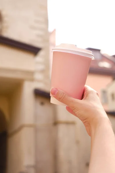 Pink paper cup with coffee in a woman\'s hand. It\'s time to drink coffee in the city. Disposable paper cup close-up on the background of the city. Empty space for text, layout. Coffee to go concept